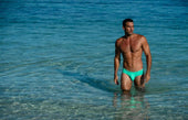 A man stands in clear shallow ocean water wearing bright turquoise men’s bikini swimwear highlighting bold beach style.