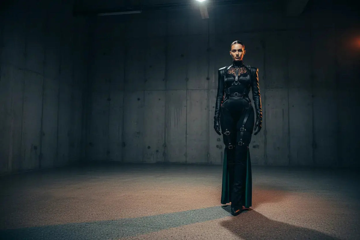 Editorial fashion shot of a figure in a black architectural harness and structured bodysuit with chrome hardware against a brutalist concrete studio backdrop