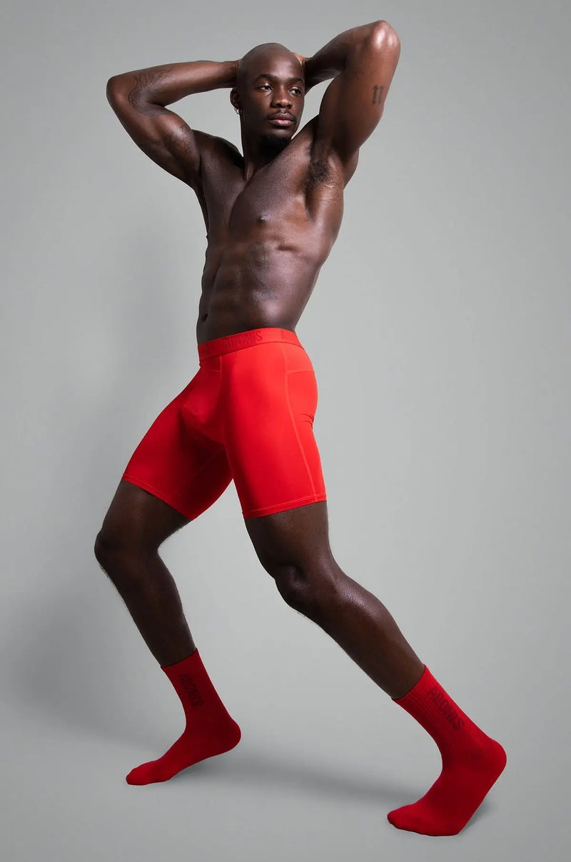Bare-chested man wearing bright red compression shorts and matching socks against grey backdrop.