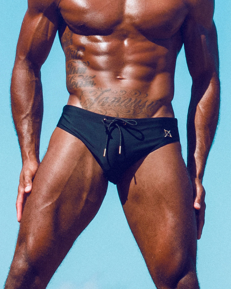 An athletic man wearing black swim briefs against a bright background.