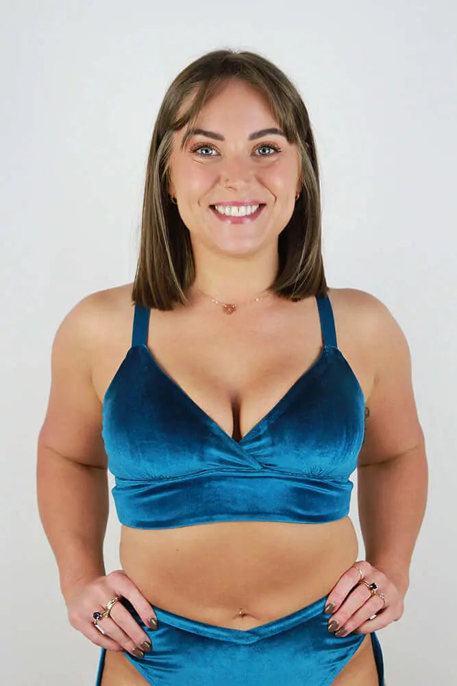 A smiling woman wearing a teal velvet bralette against a plain white background.