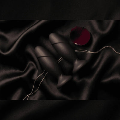 Anal Adventures black silicone anal plugs on black satin with rose petal, dark luxury editorial flat lay
