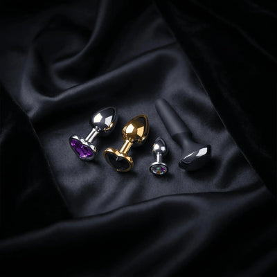 Premium anal plug collection — silver, gold and black silicone plugs with jewelled bases arranged on dark satin