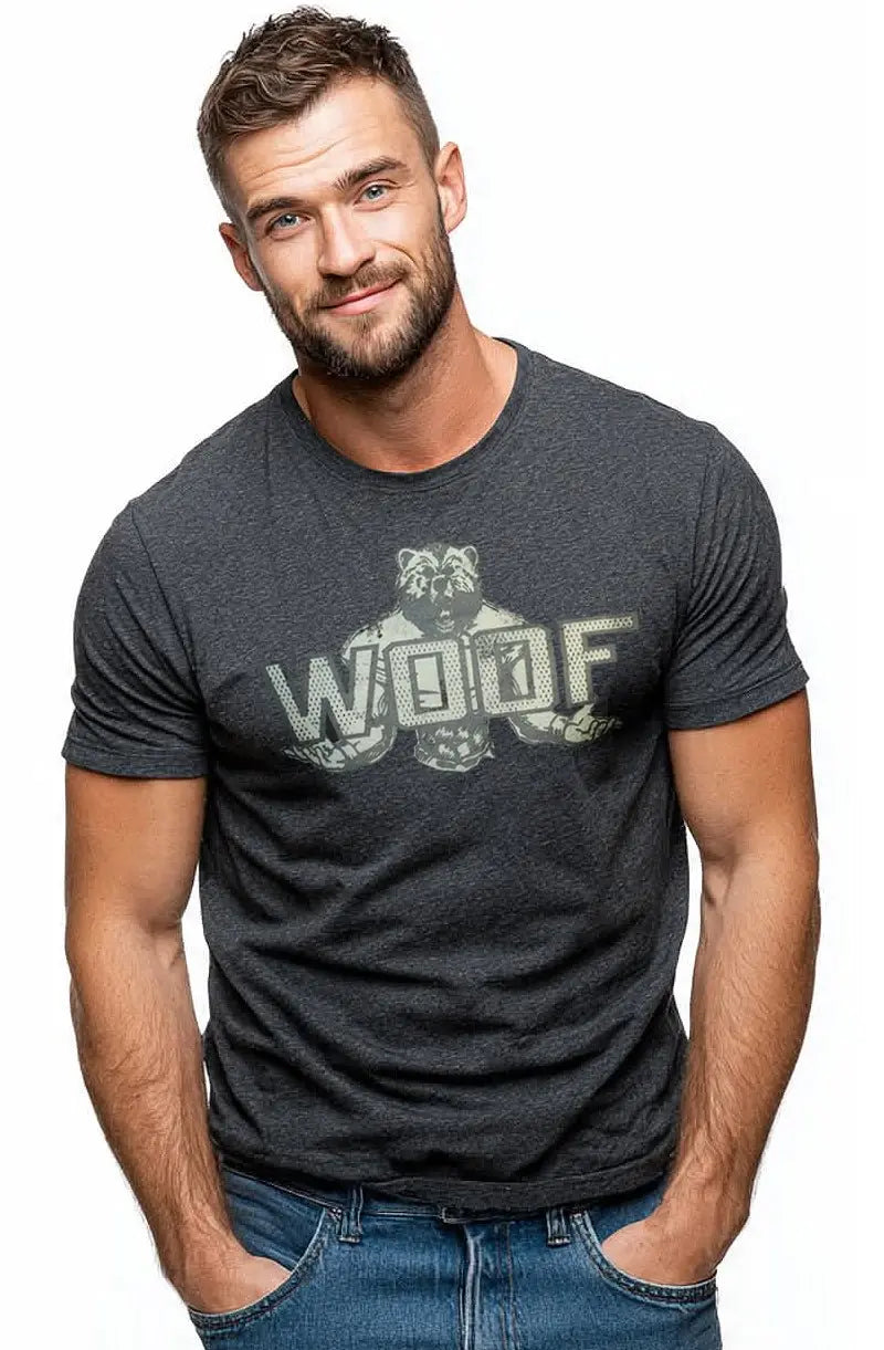 A bearded man wears grey graphic t-shirt and blue jeans, standing against white.