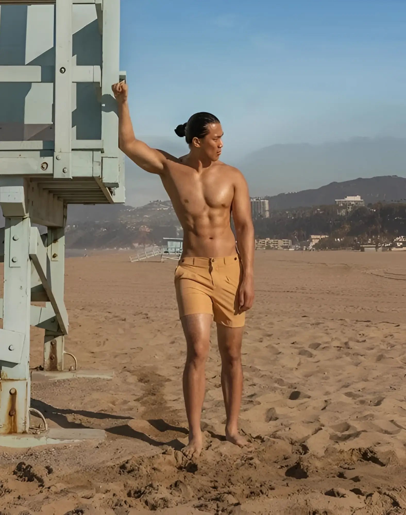 A man stands on a sandy beach wearing 2EROS Avalon beige shorts with belt loops and button closure