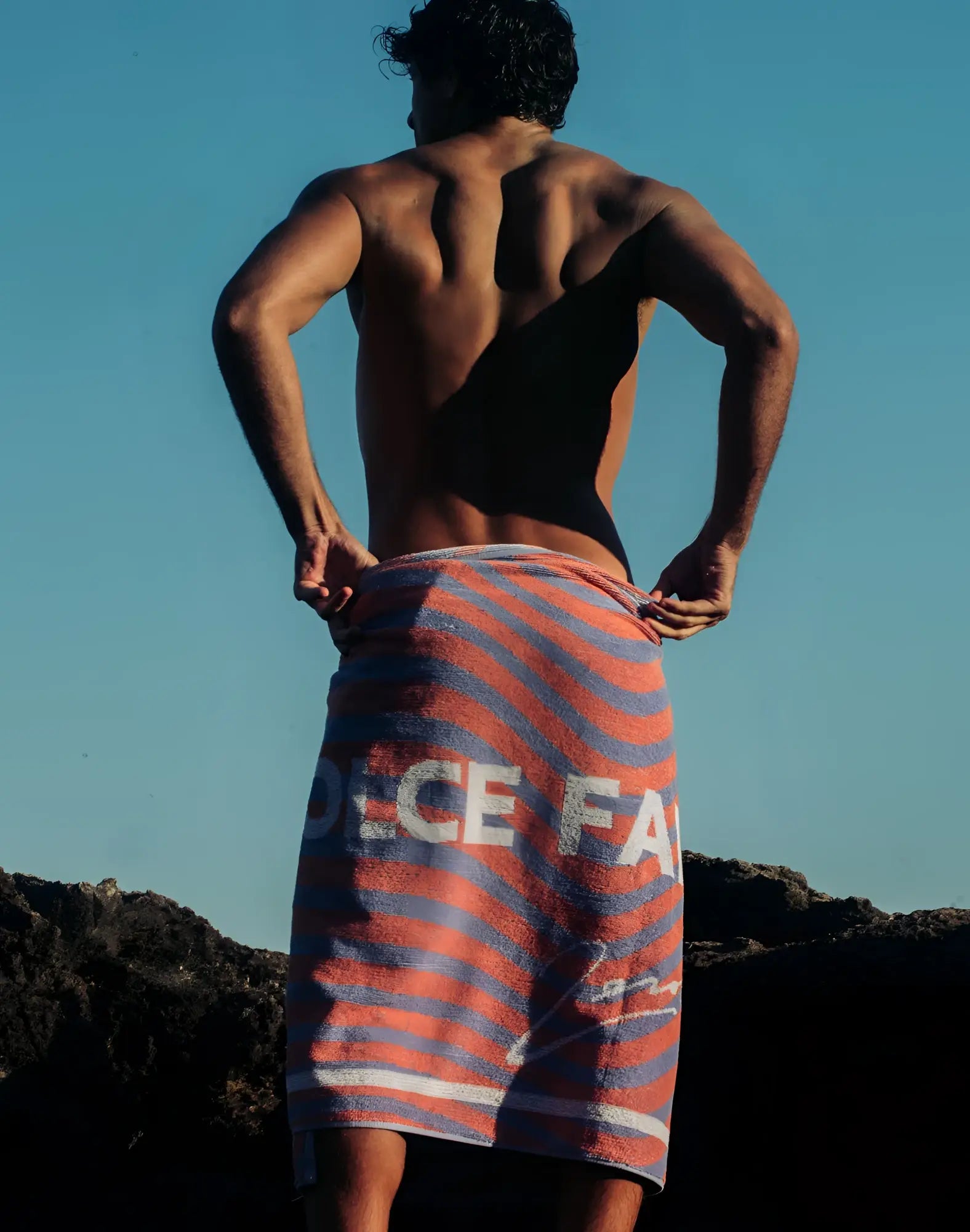 A vibrant orange and blue striped 2eros beach towel with bold white dolce and gabbana lettering wrapped around a man’s waist