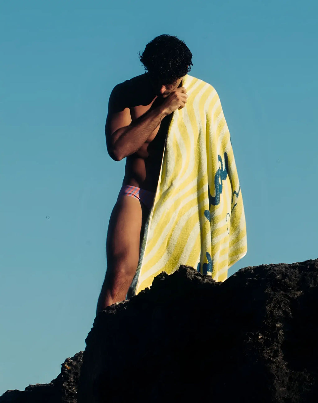 A vibrant yellow 2EROS beach towel with bold white stripes and blue script logo wrapped around a mans shoulders