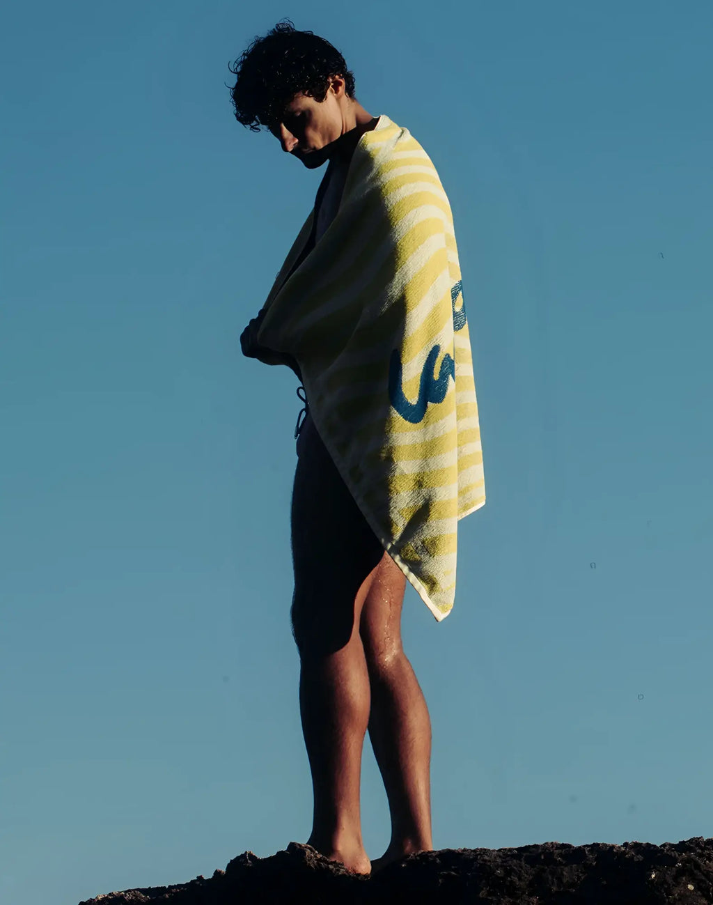 A 2EROS beach towel in yellow and white stripes with a blue logo draped over a person’s shoulders