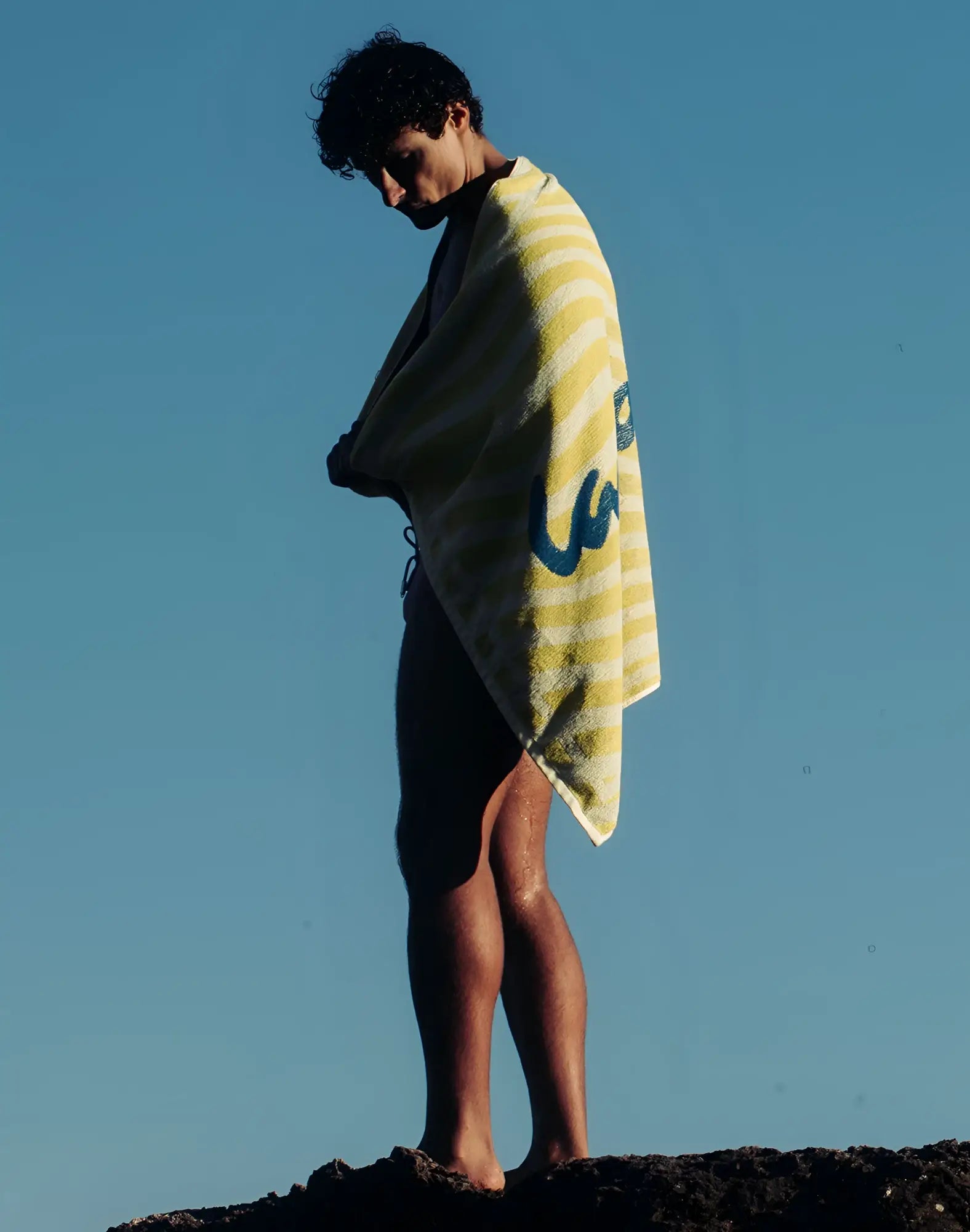 A 2EROS beach towel in yellow and white stripes with a blue logo draped over a person’s shoulders