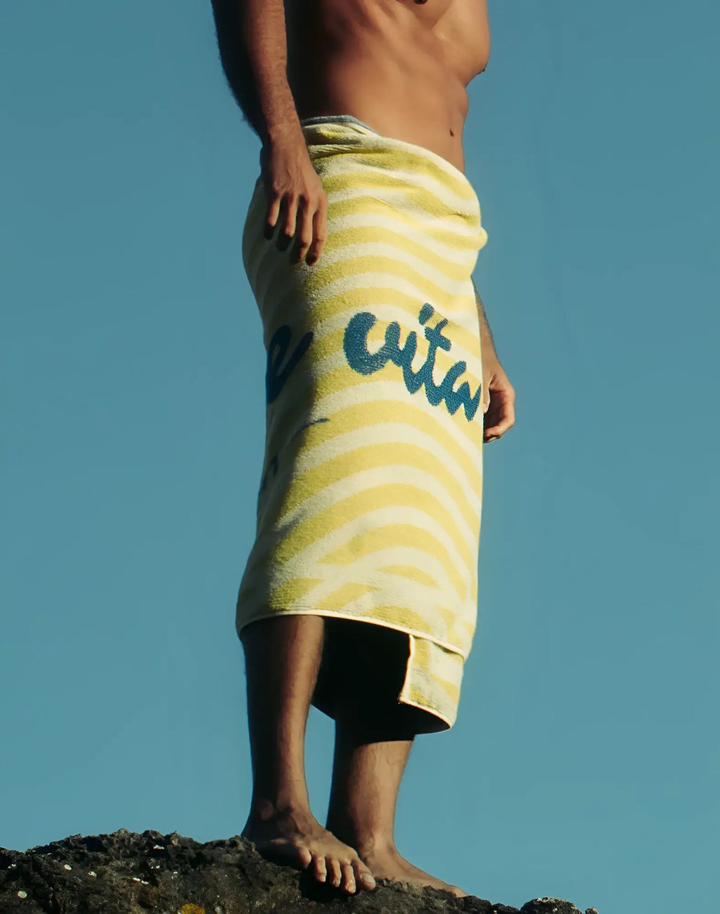 A yellow and white striped 2EROS beach towel with blue cursive lettering wrapped around a person’s waist