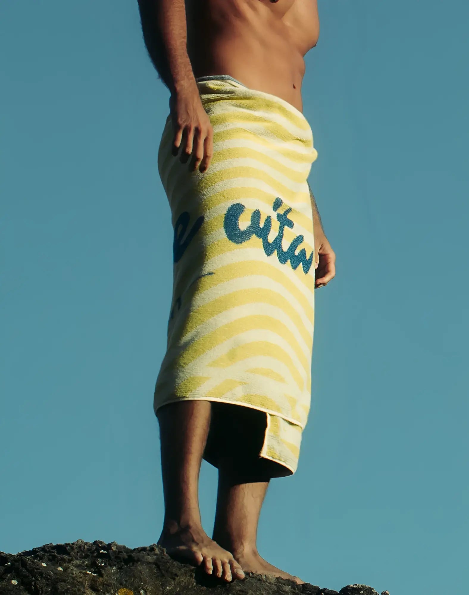 A yellow and white striped 2EROS beach towel with blue cursive lettering wrapped around a person’s waist