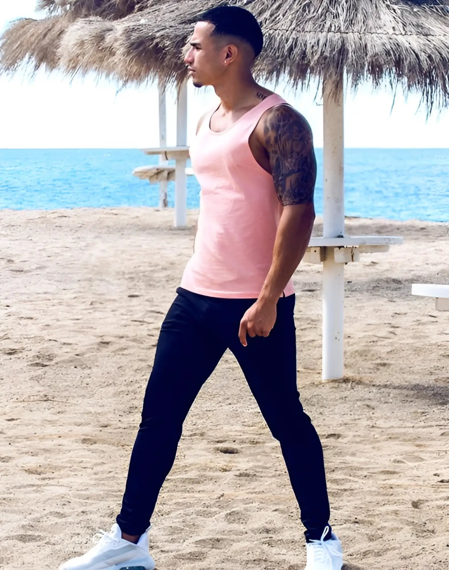A man wearing black 2EROS P60 Bondi pants stands on the beach in a pink sleeveless tank top