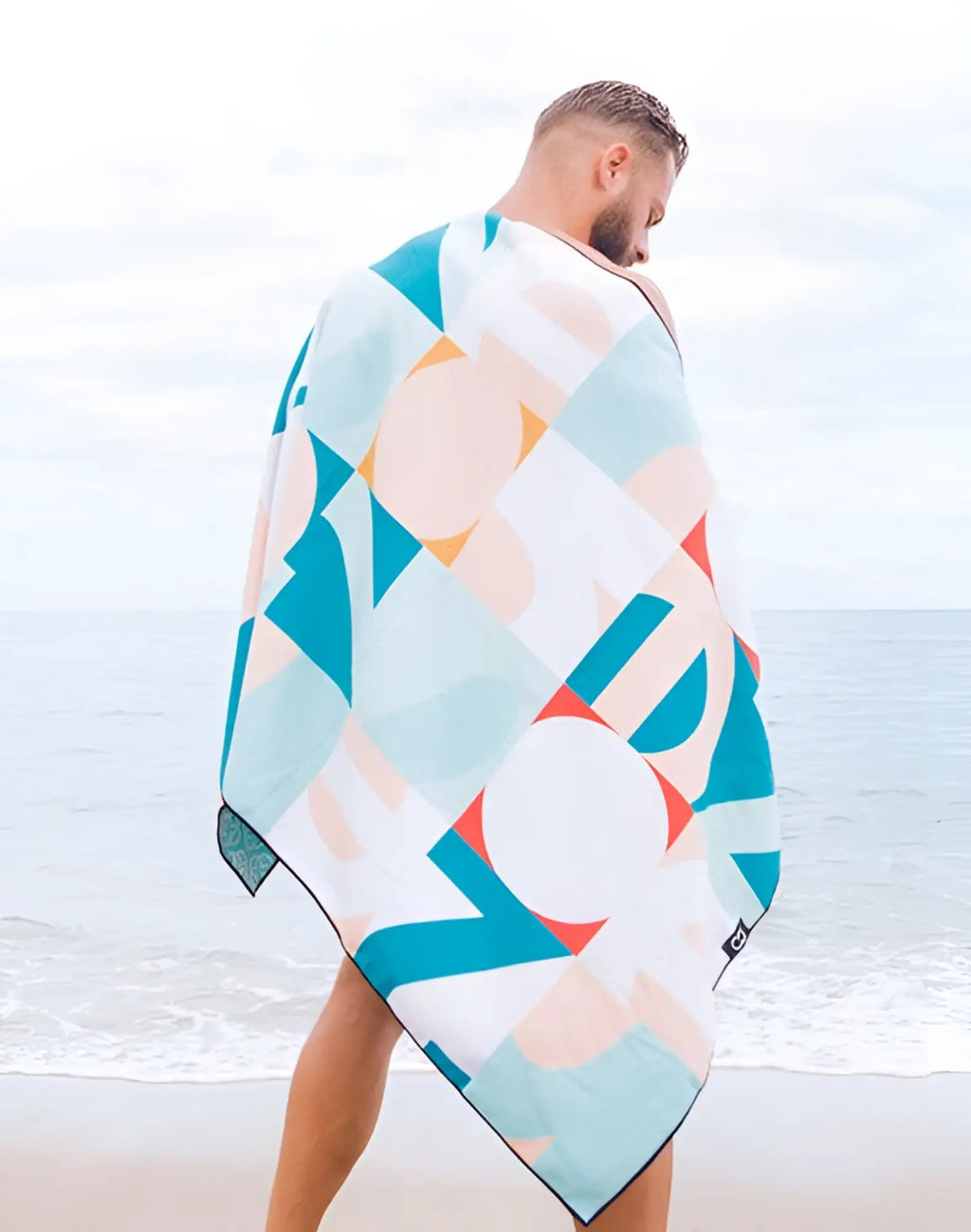 A man wearing a 2EROS sand-free beach towel in insignia turquoise with vibrant teal coral and white geometric patterns