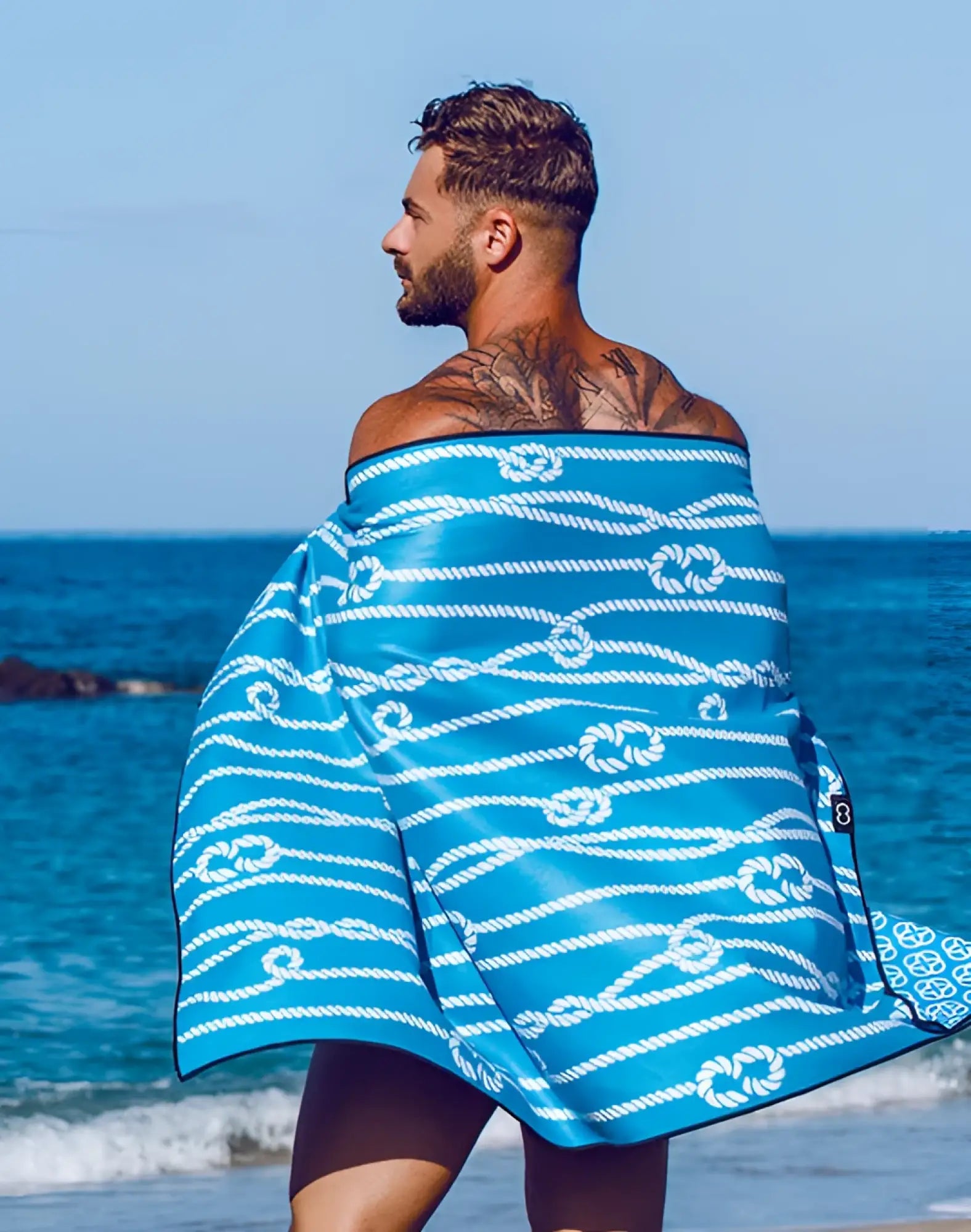 2eros sand-free beach towel in manrope blue with white rope knot pattern wrapped around a mans shoulders front view
