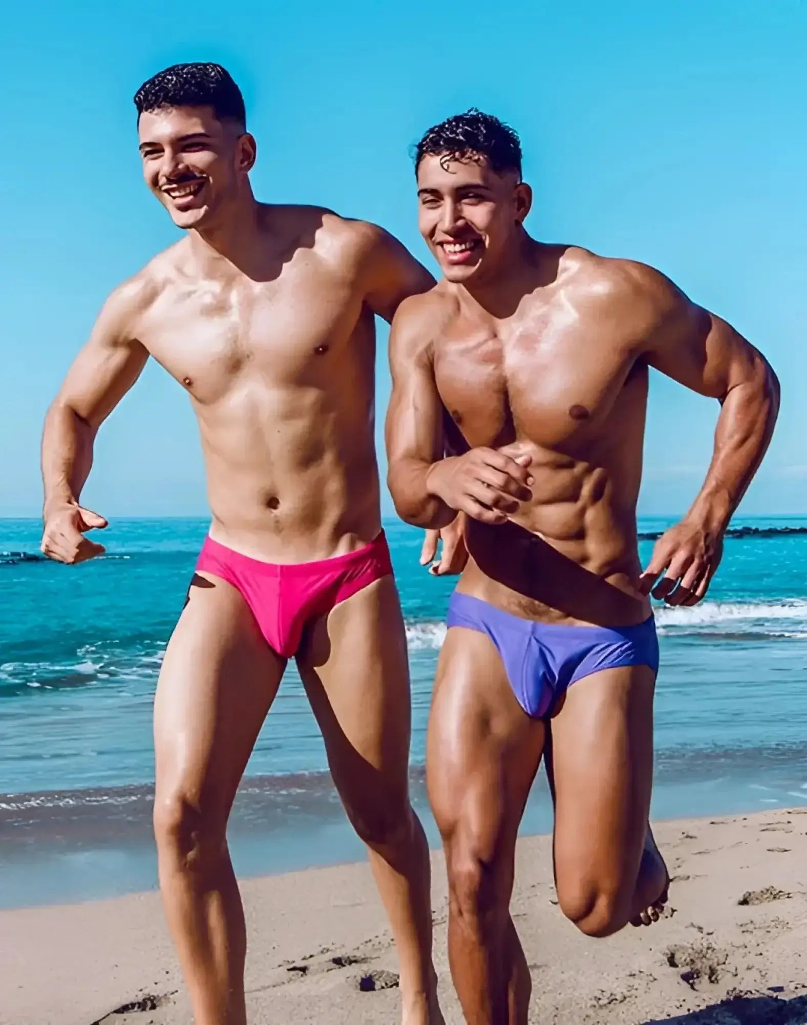 Vibrant fuchsia red 2eros v10 core swim trunks worn by two muscular men on a sunny beach in pink and blue colors