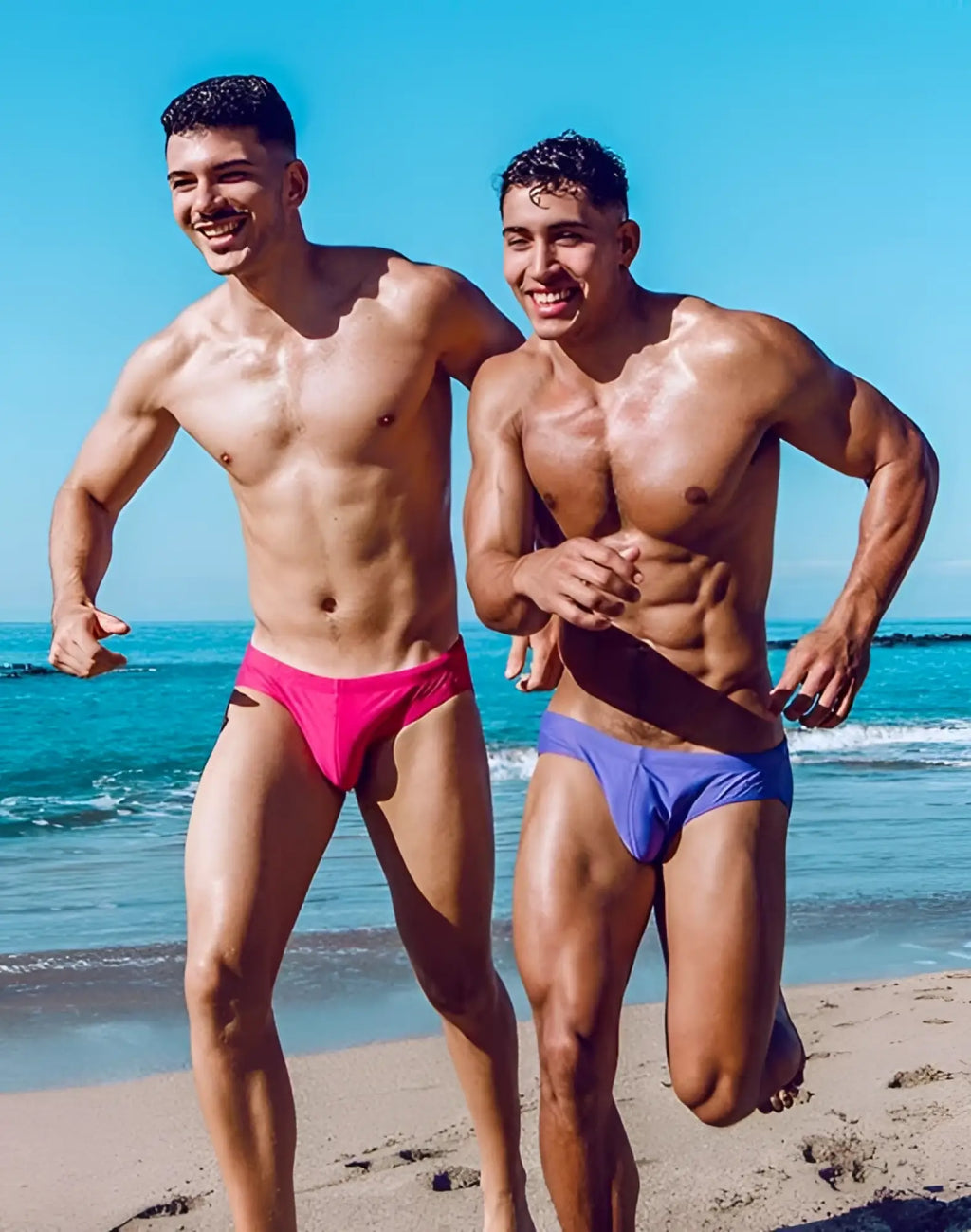 Vibrant fuchsia red 2eros v10 core swim trunks worn by two muscular men on a sunny beach in pink and blue colors