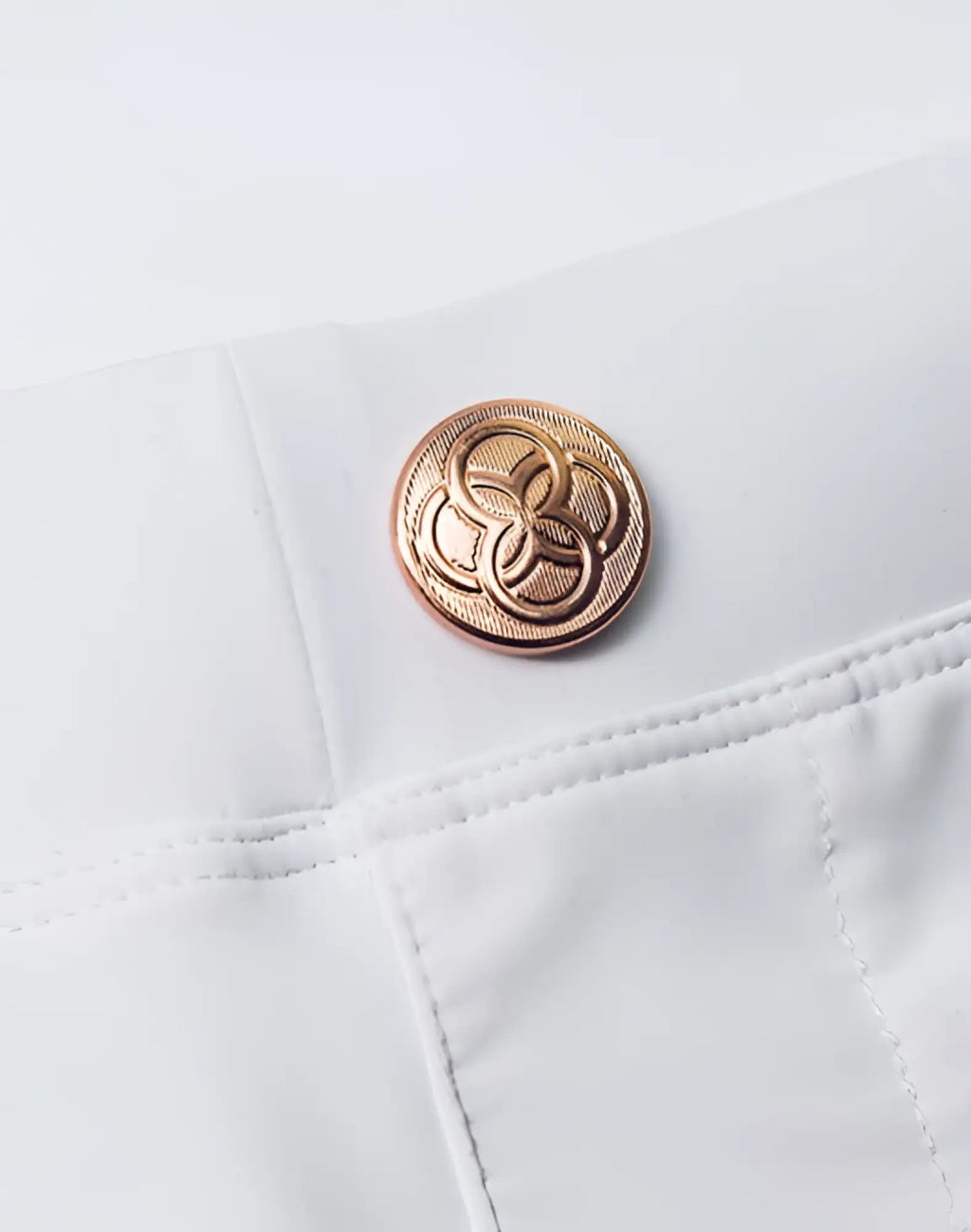 A rose gold button with an intricate interlocking circle design on white fabric of 2eros v60 bondi swim trunks