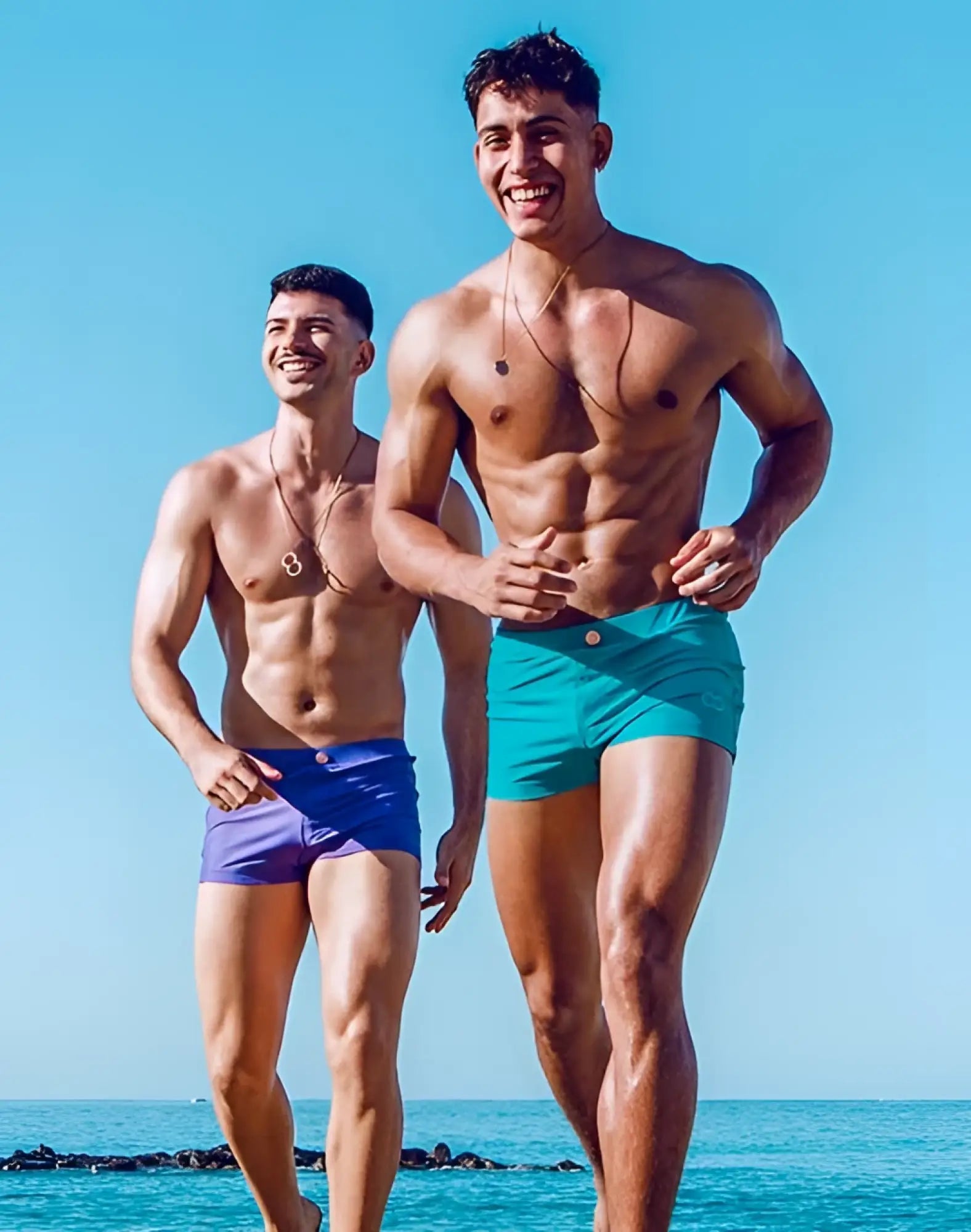 Two muscular men wearing 2eros v60 bondi series 2 swim trunks in dusted purple and vibrant turquoise stand on a beach