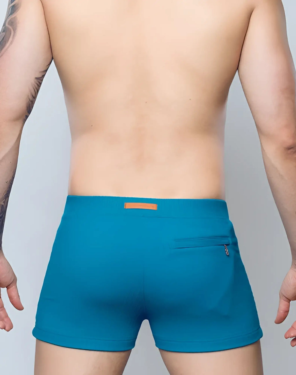 Back view of 2EROS V60 swim trunks in railroad teal with textured fabric, orange waistband label, and zippered back pocket