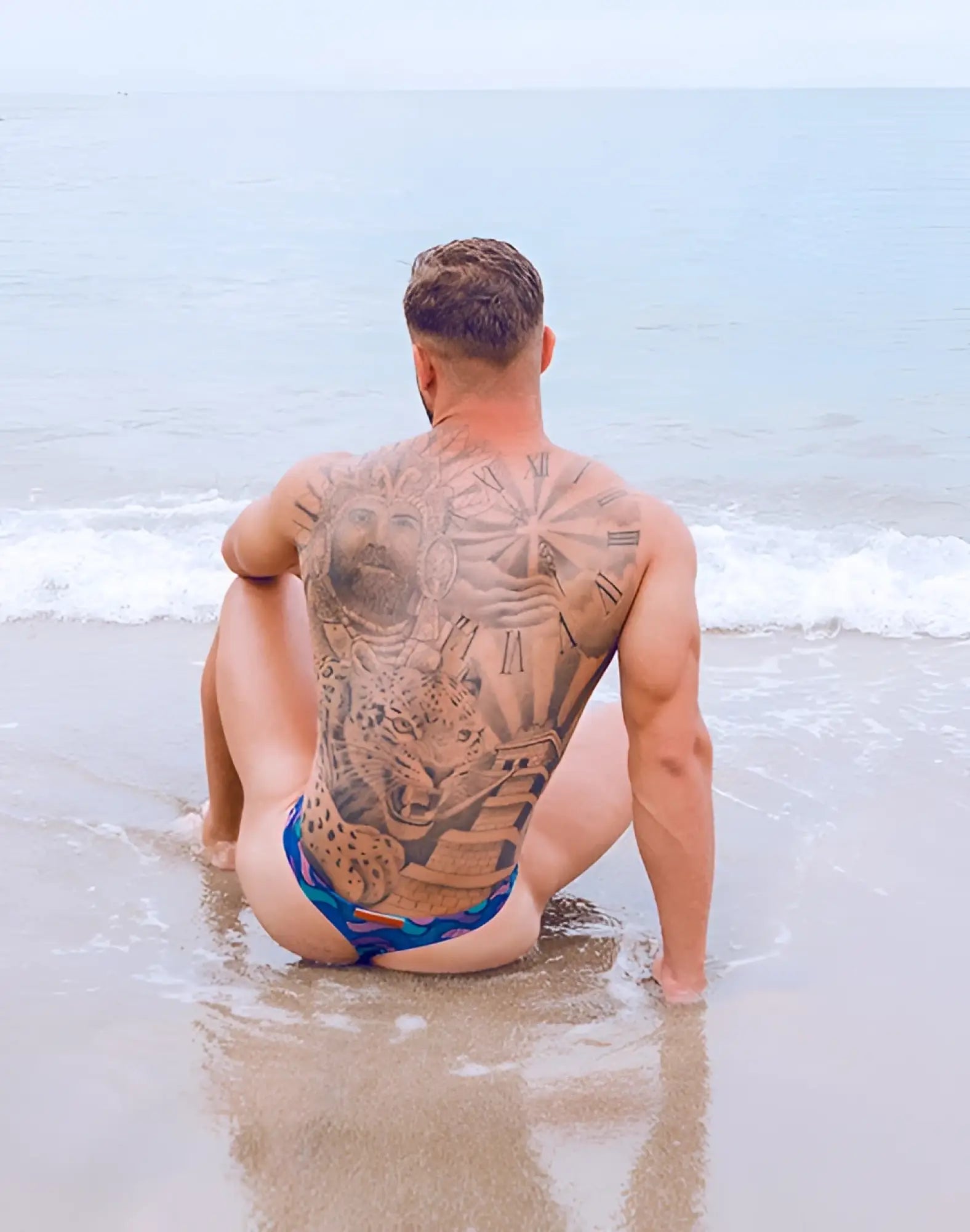 A man sitting on a beach wearing 2EROS V80 print swim thong in ocean blue with intricate orange designs