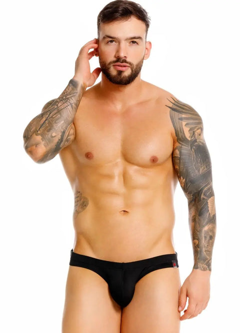 Bearded man with arm tattoos in black briefs against a white background.