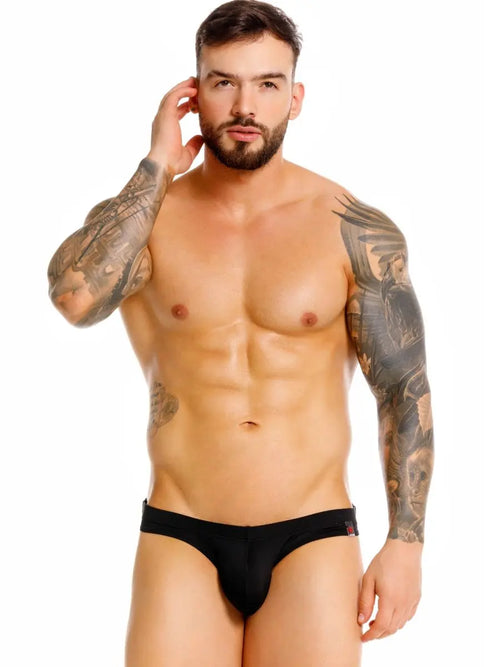Bearded man with arm tattoos in black briefs, hand to head, on white.