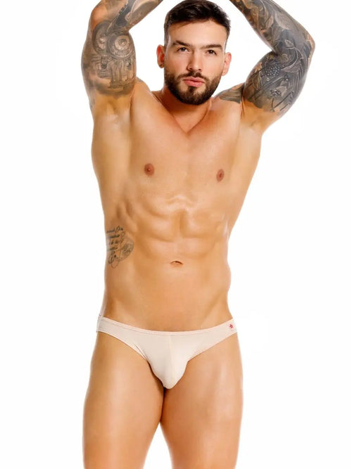 Bearded man with arm tattoos in light briefs, arms raised, white background.