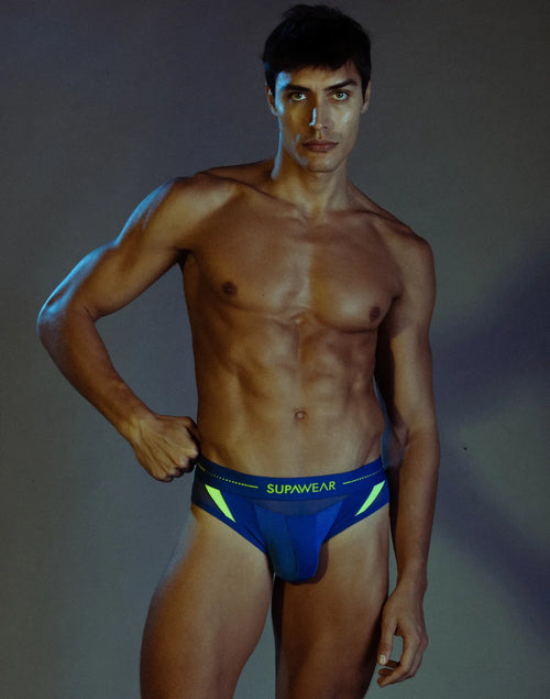 Man wearing SUPAWEAR blue brief with neon accents, hand on waistband.