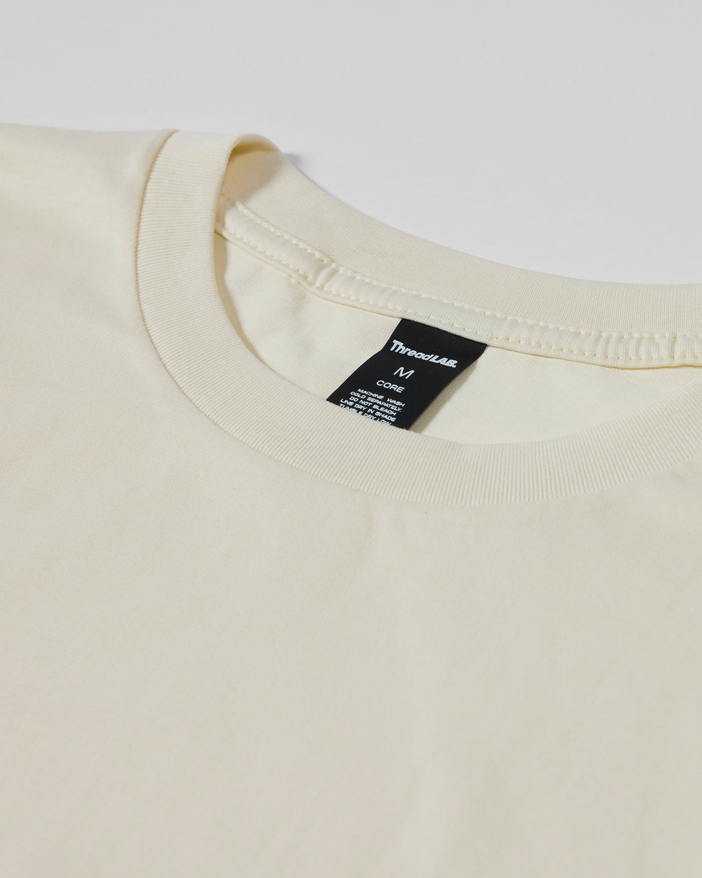Close-up of a cream-colored crew neck T-shirt with a black ThreadLab brand tag at the neckline.