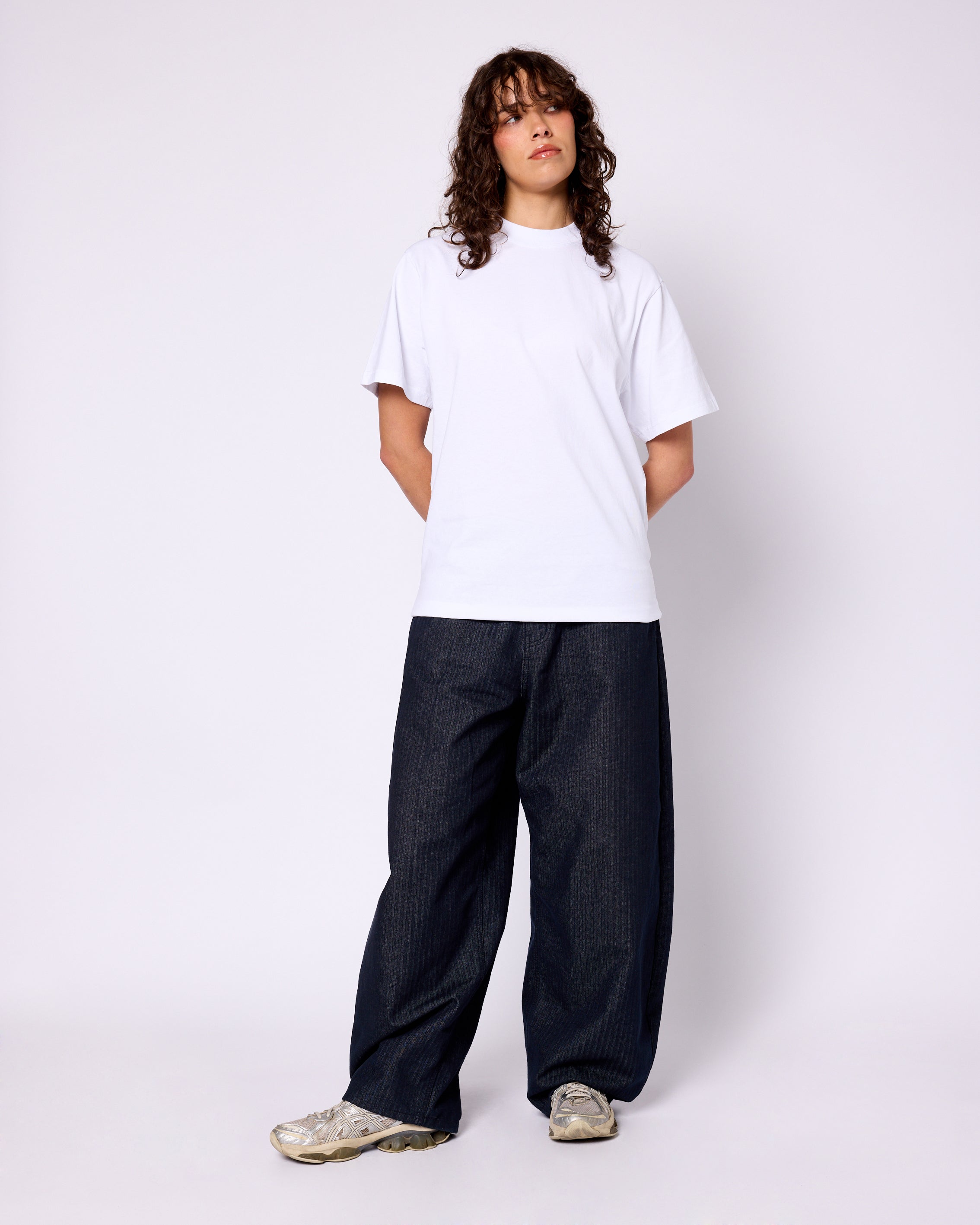 Model wears a clean white crewneck tee with a boxy, relaxed fit, paired with dark, wide-leg pinstripe denim pants.