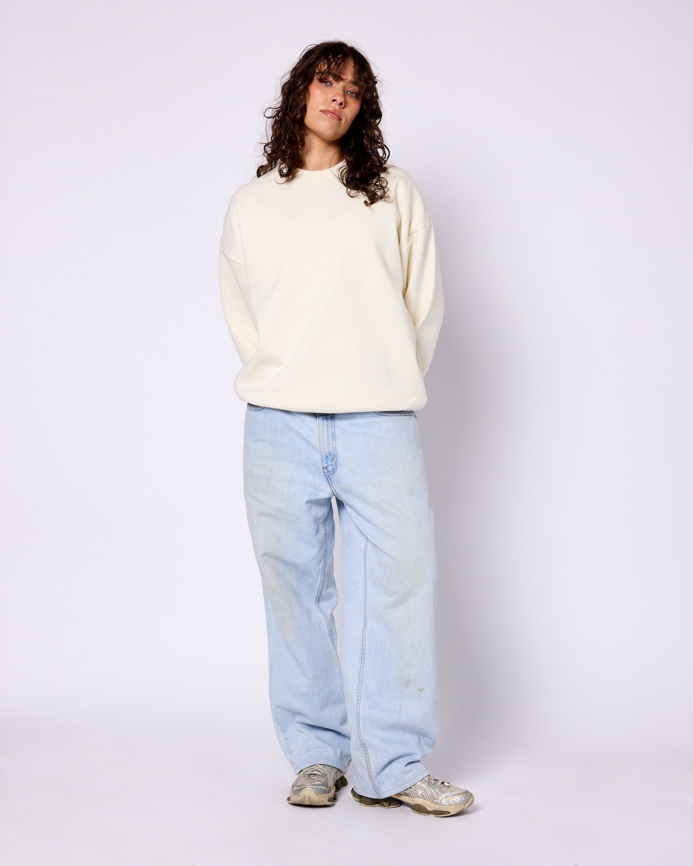 Model wearing a cream-colored, long-sleeve crewneck sweatshirt with dropped shoulders and a relaxed fit.