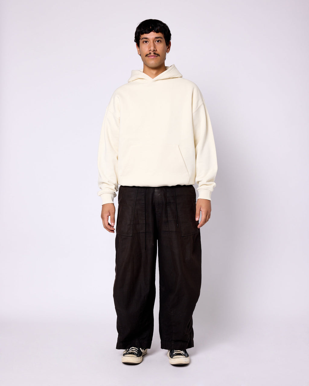 A man wears a plain, off-white, oversized cotton hoodie with a front kangaroo pocket and rib-knit cuffs.