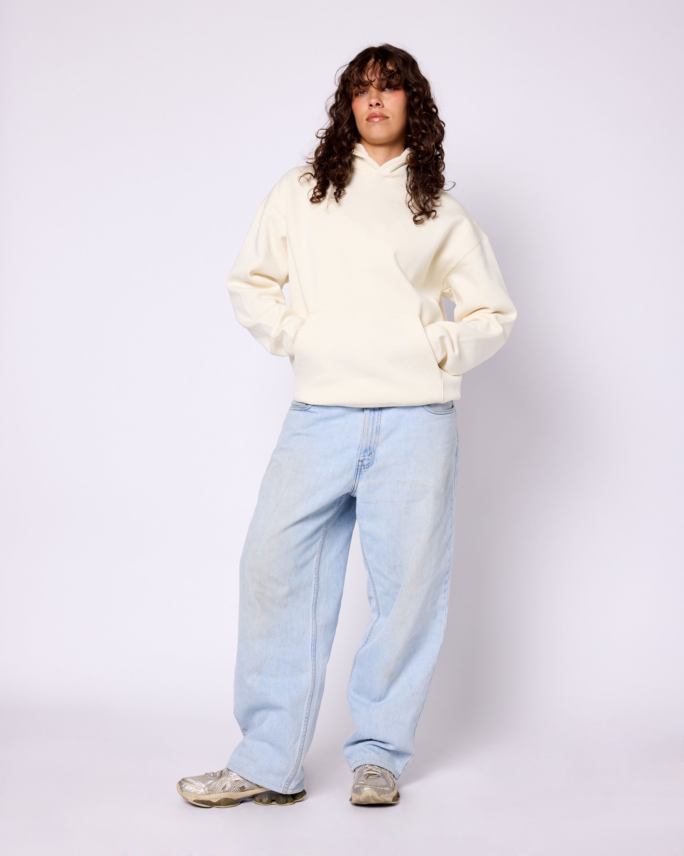 Model wearing an oversized, cream-colored Core Hoodie with a front kangaroo pocket, paired with light-wash baggy jeans.