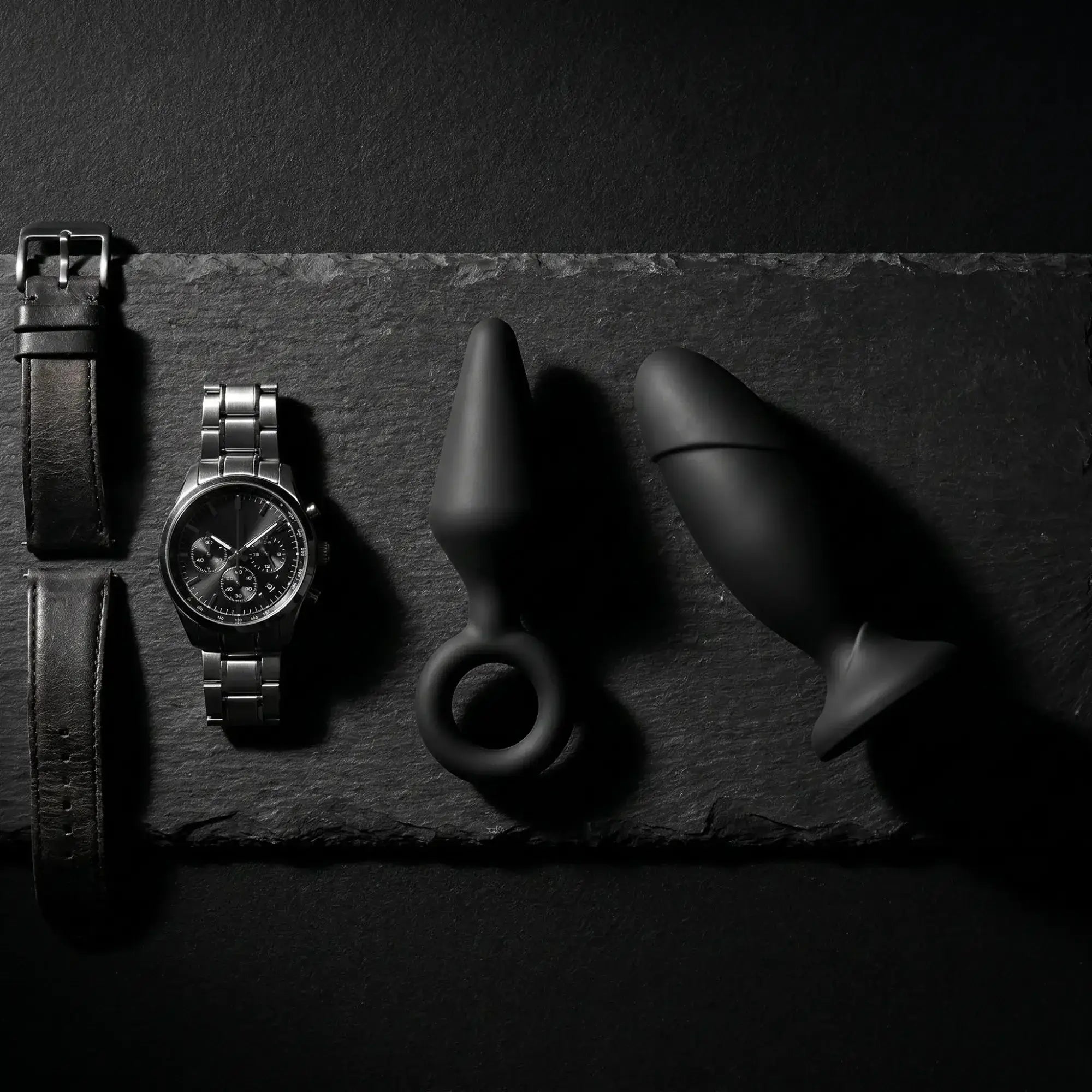 Anal Adventures black silicone anal plugs on dark slate with leather strap, masculine luxury editorial flat lay