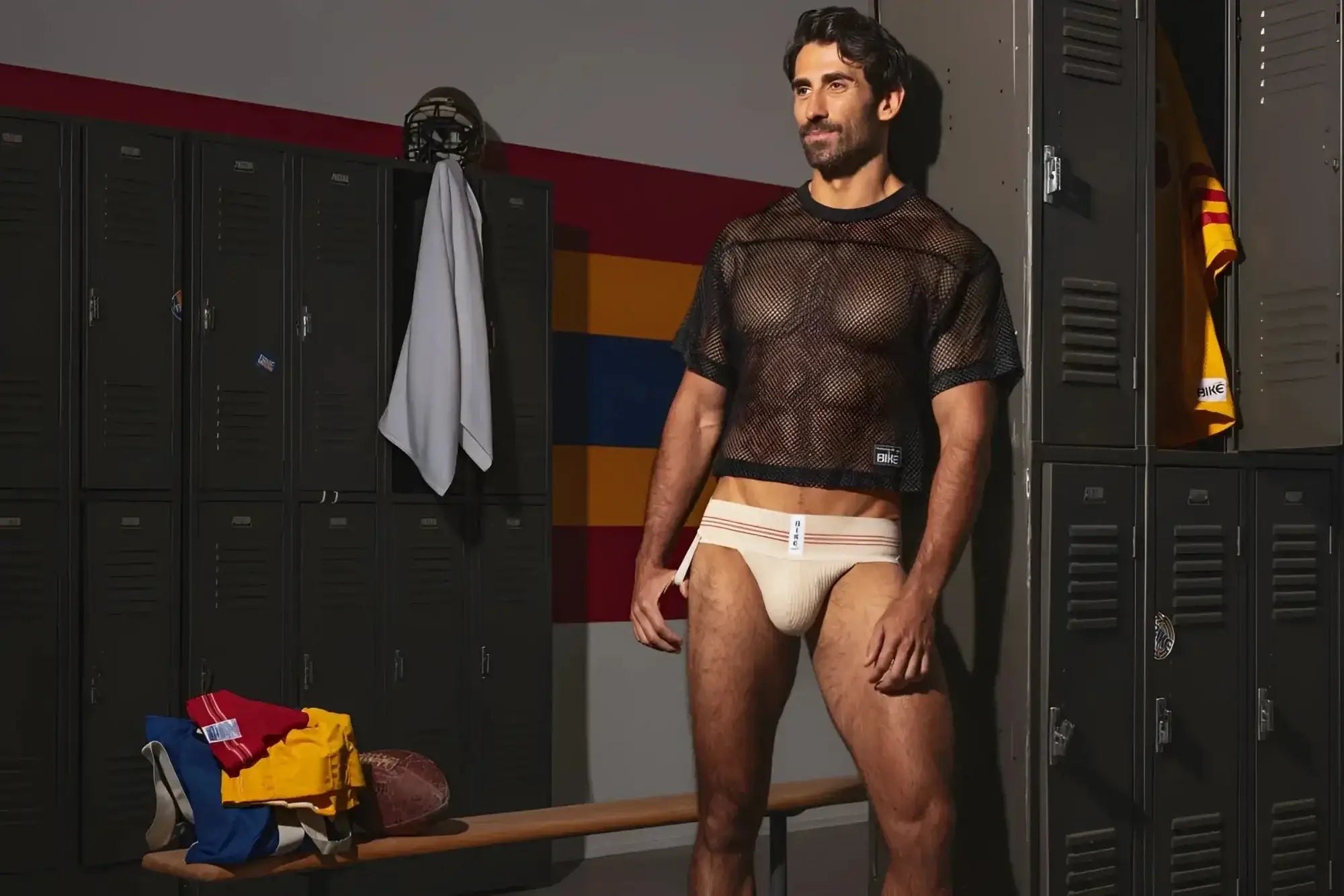 Lifestyle shot: A model wears the BIKE Athletic BAS333 Classic Retro jockstrap in vintage cream in a locker room.