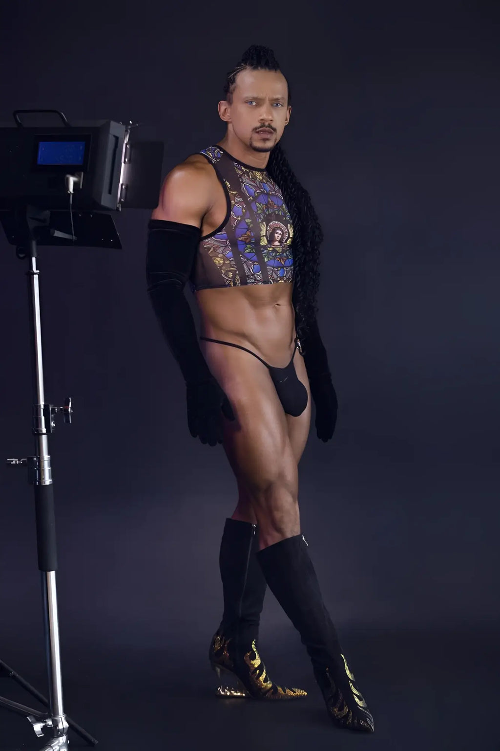 A man wearing a black and gold patterned CandyMan 99805 stained glass tank top with sheer mesh and matching thigh-high boots