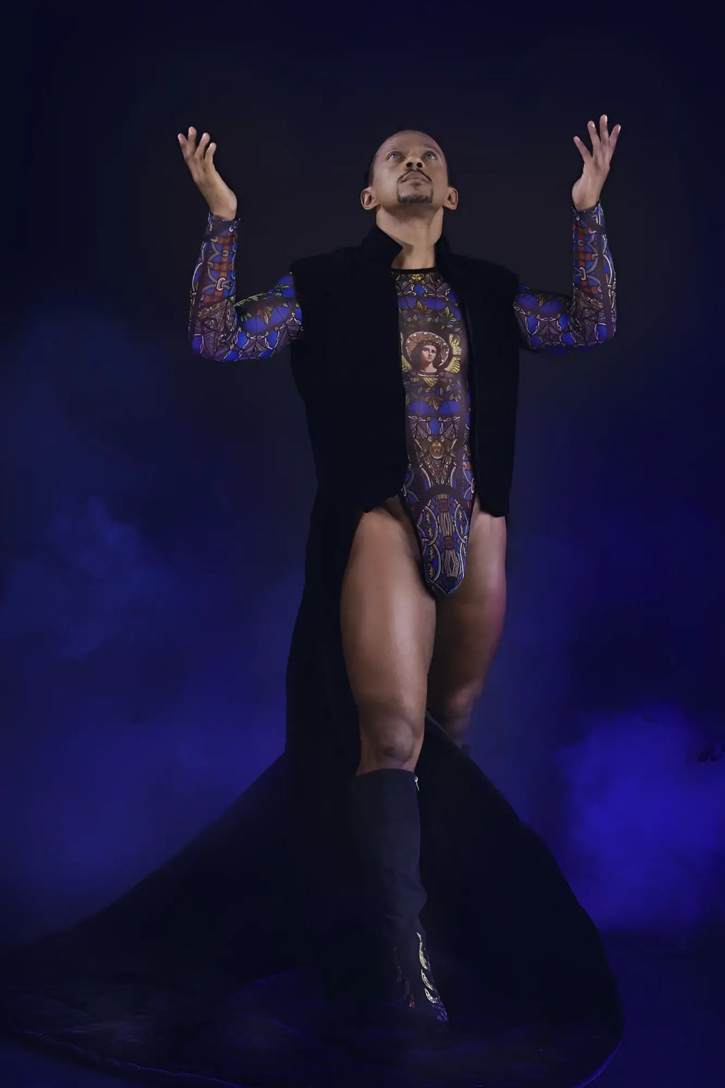 A man wearing a black candyMan 99814 stained glass bodysuit with blue and gold ornate patterns and a black vest