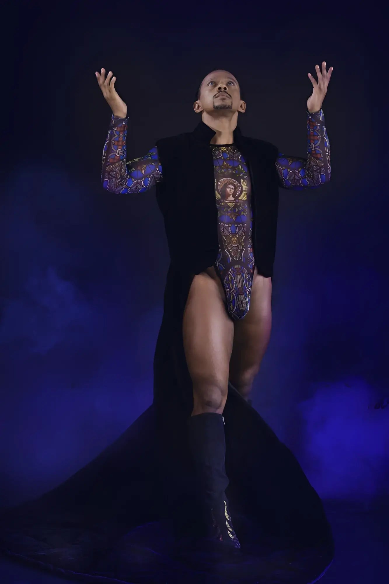 A man wearing a black candyMan 99814 stained glass bodysuit with blue and gold ornate patterns and a black vest