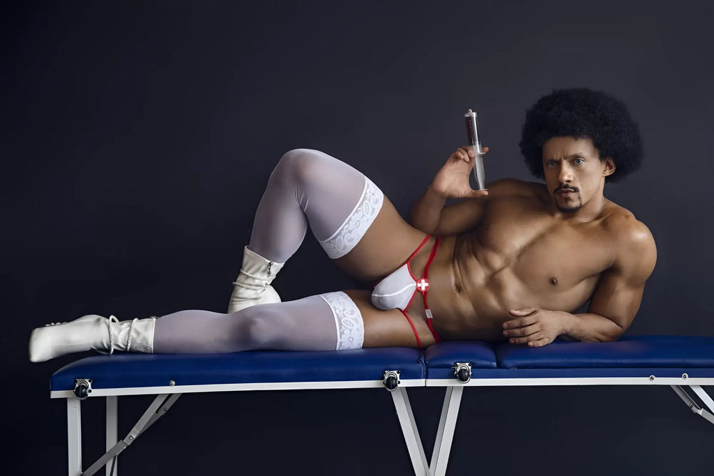 A muscular man with an afro lies on a blue table wearing candyMan 99833 red-white medical-themed murse thongs and white lace stockings