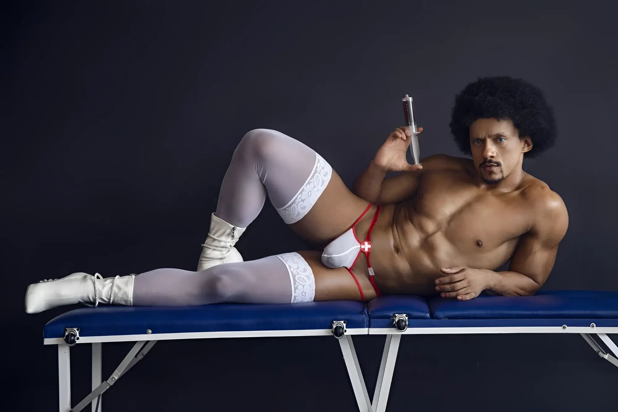 A muscular man with an afro lies on a blue table wearing candyMan 99833 red-white medical-themed murse thongs and white lace stockings
