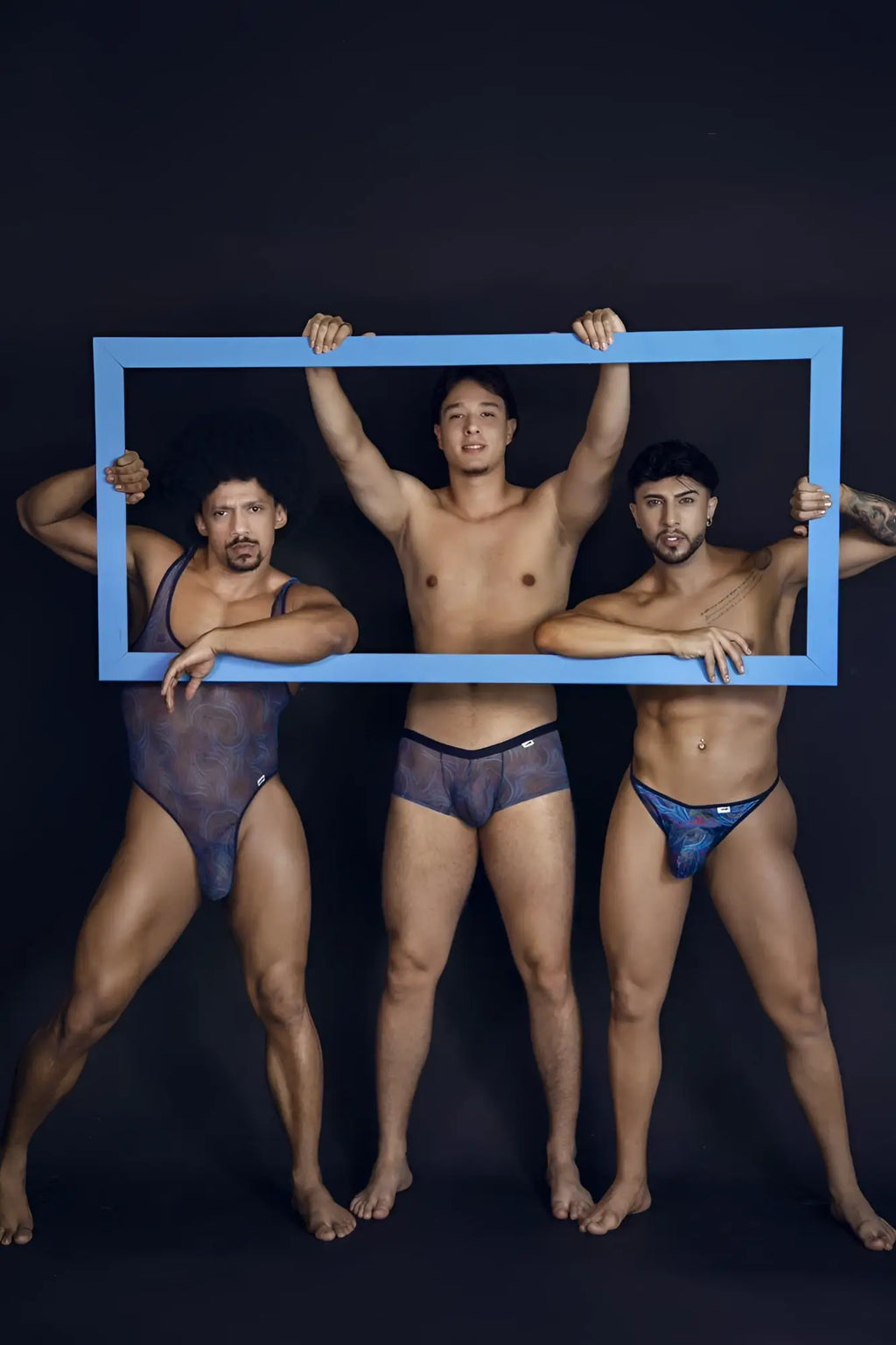 Three men holding a blue rectangular frame showcasing candyman 99875 seawater thongs with multicolor print