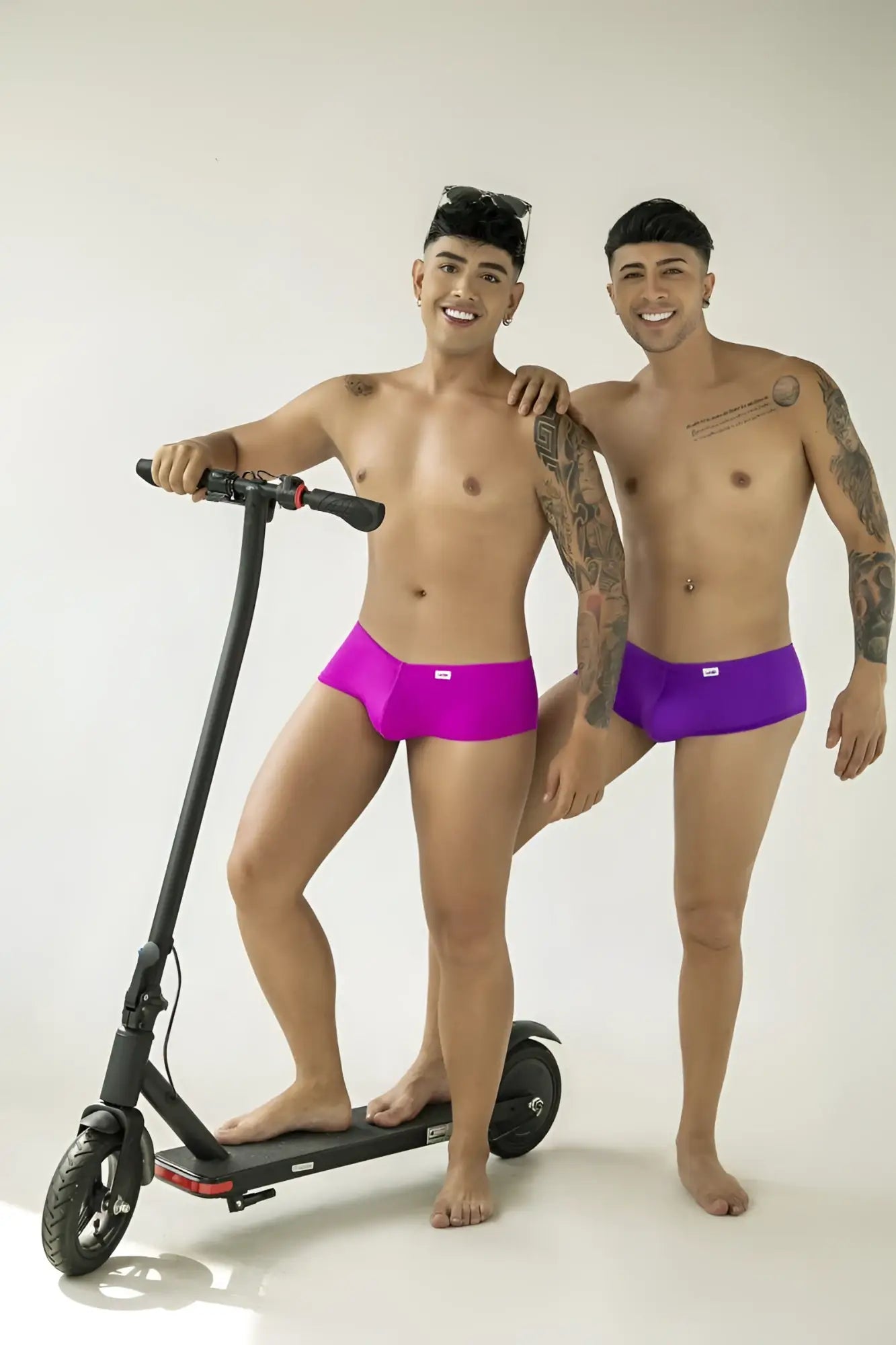 Two models wearing CandyMan magenta and purple low-rise trunks, standing with a black electric scooter.
