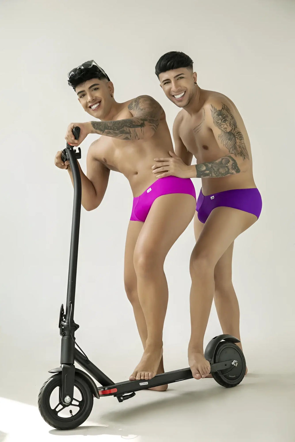 Two shirtless men wearing Orchid-colored CandyMan trunks standing on a black electric scooter against a white background.