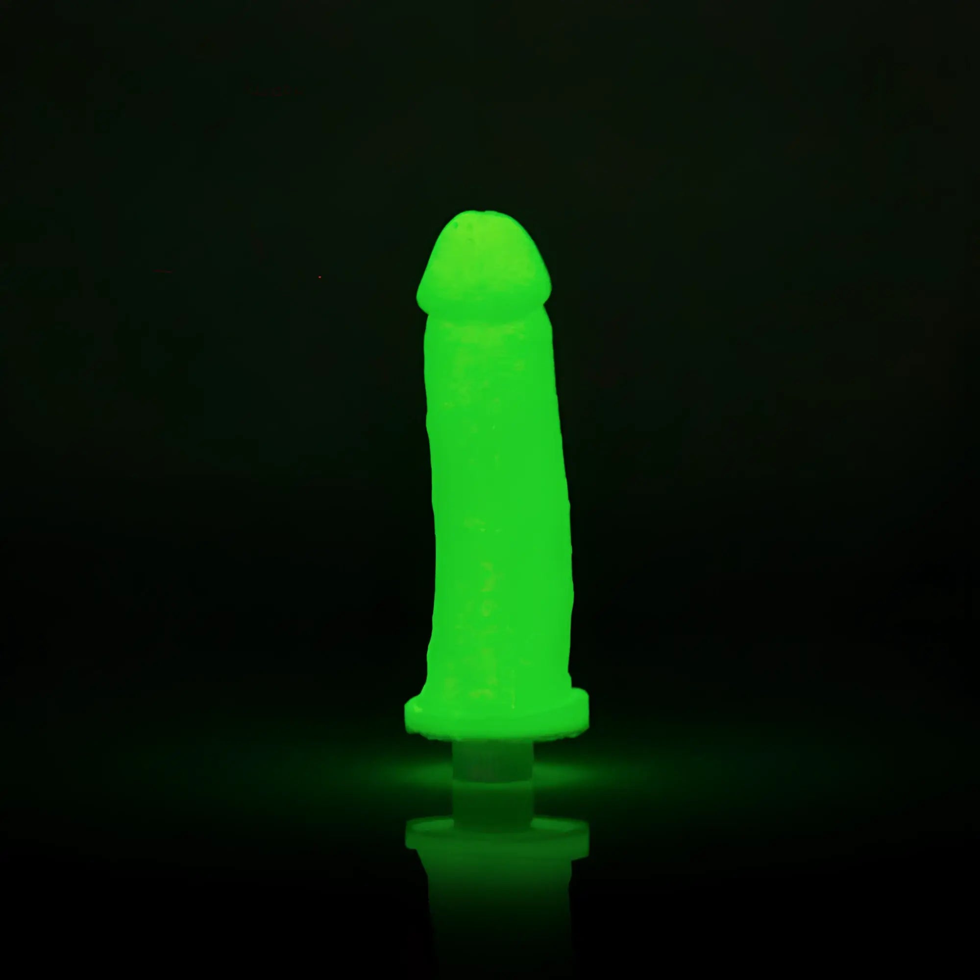 A glowing green silicone toy clone a willy with a rounded head and tapered base standing upright on a reflective surface