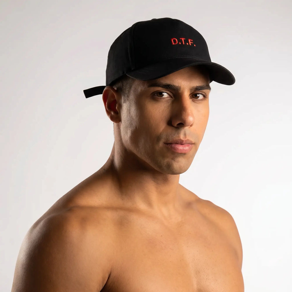Athletic man in sleek black baseball cap with white embroidered text, shirtless editorial lifestyle shot on grey backdrop