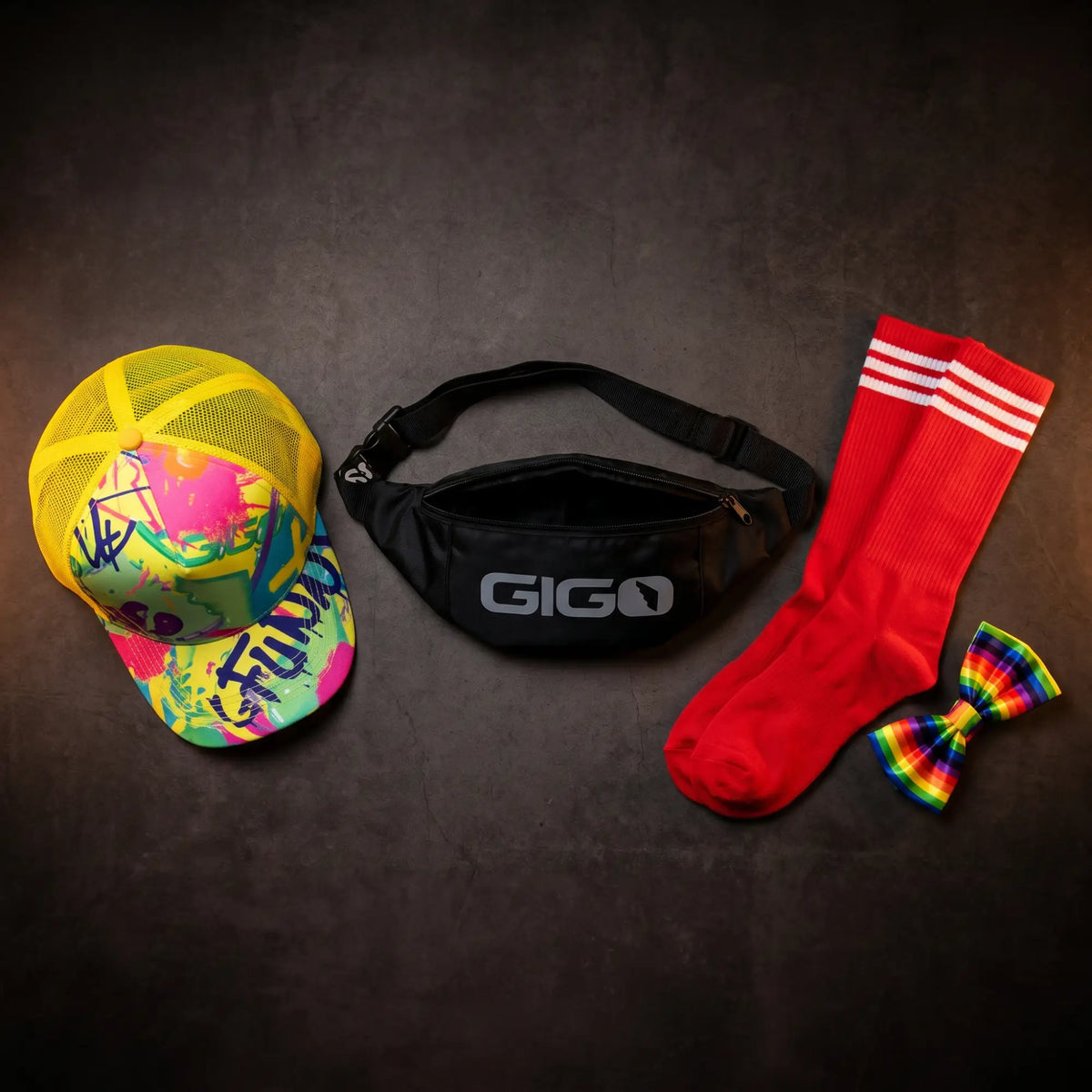 Men's accessories banner flat lay: neon yellow cap, black waistpack, red knee-high socks, and rainbow bow tie on dark surface