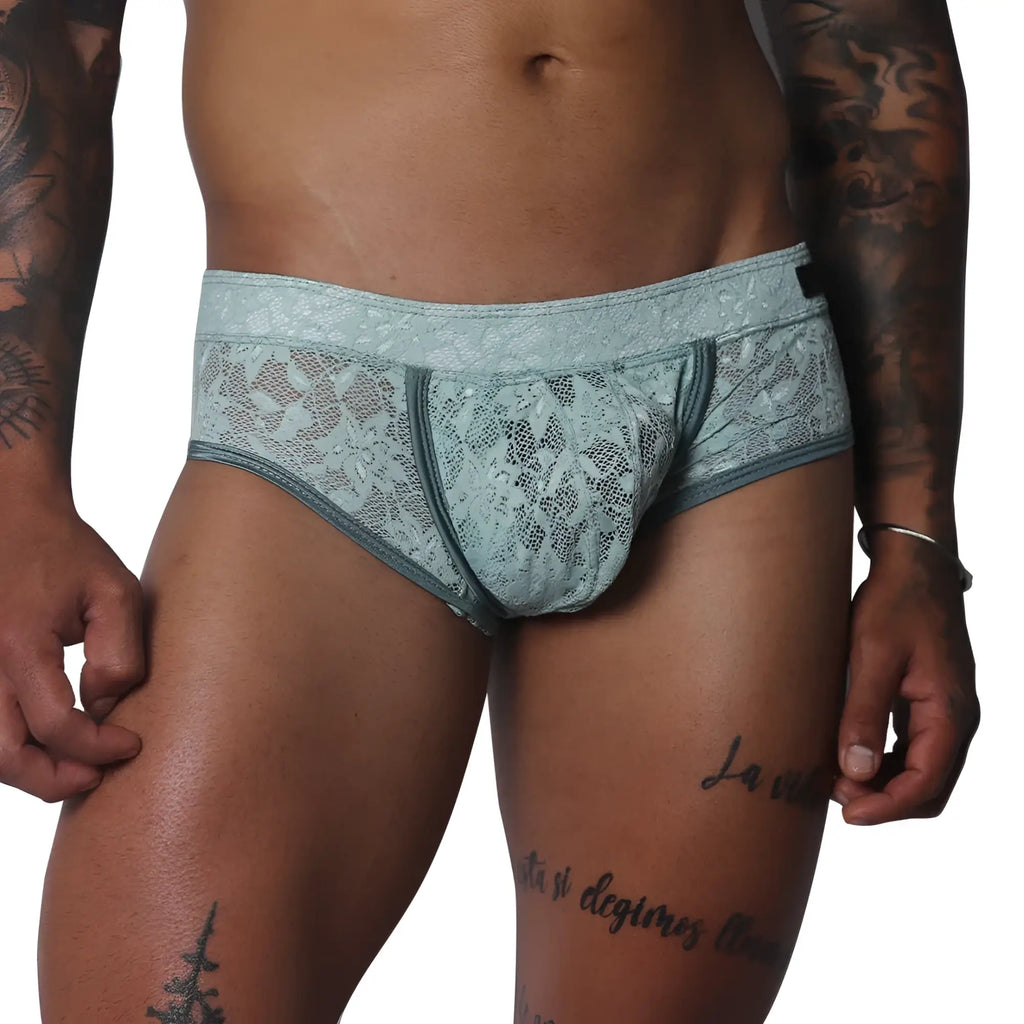 Aqua blue floral lace trunk with solid piping and a contoured pouch, modeled on a tattooed torso.