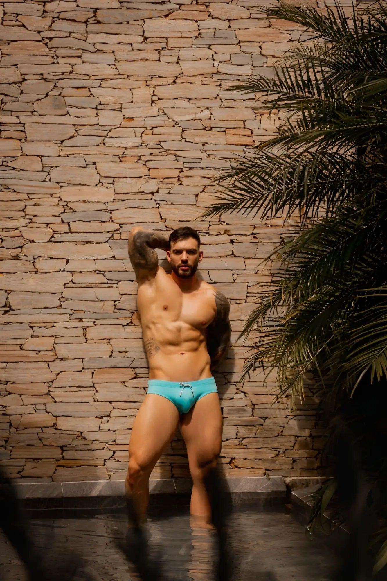 Lifestyle shot: A fit man posing in turquoise JOR 2387 Travel swim briefs against a stone wall.