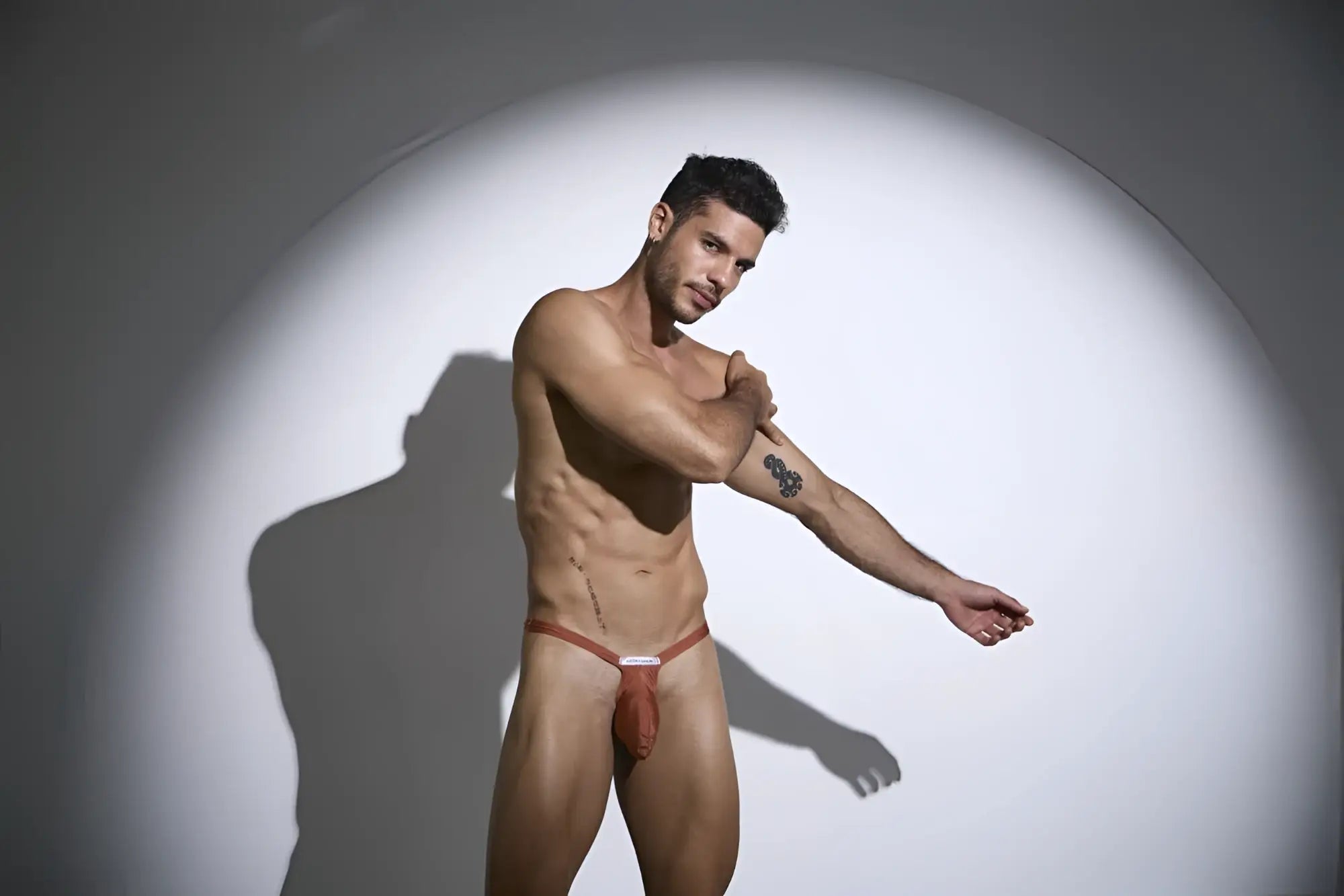 A man wearing a reddish-brown ochre justin simon xsjbu02 bulge thong jockstrap with thin straps and a white label at front