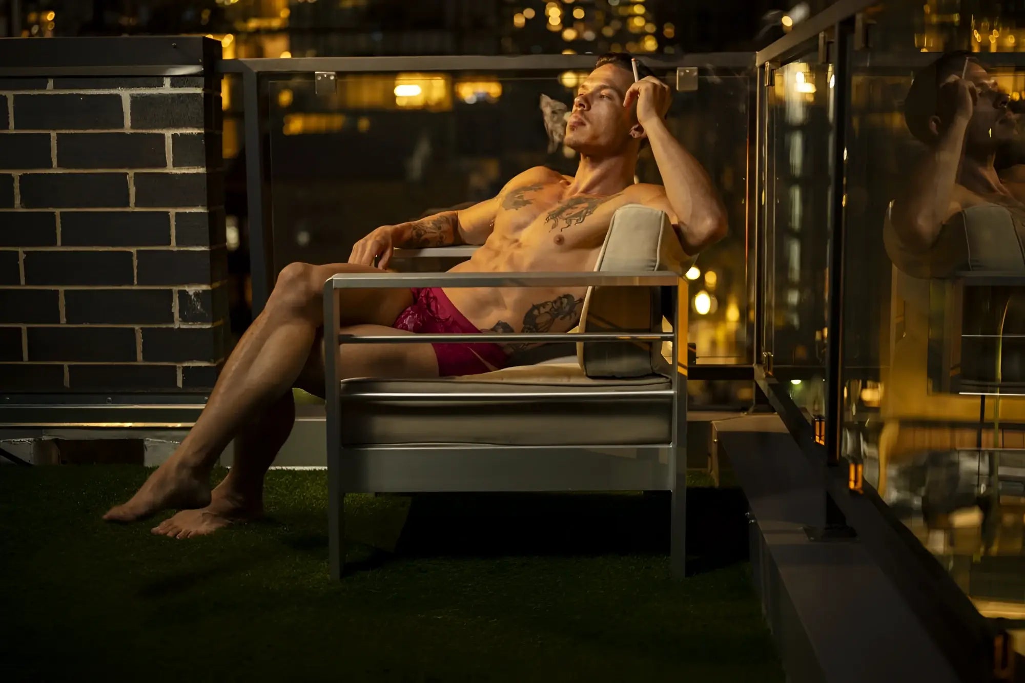 A man with tattoos lounges in a modern metal-framed chair wearing male power 126-289 lucifer cut out shorts in burgundy